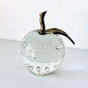 May include: A clear glass apple paperweight with internal bubbles. It has a gold-colored stem and leaf. The apple is sitting on a white surface. Decorative home decor.