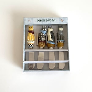 May include: Set of four cat-shaped butter knives in a box. The knives are decorated with different cat designs and have stainless steel blades. The box is white with blue trim and the words "Debbie Mumm" printed on the front.