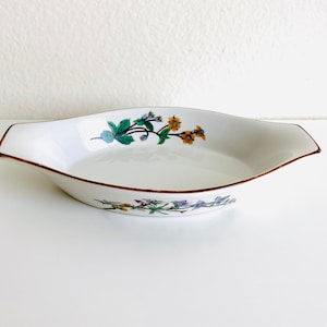 Puede incluir: Plato para hornear ovalado de cerámica blanca con un diseño floral en tonos verdes, amarillos y azules. El plato tiene un borde dorado.