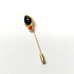 Peut inclure: Une broche dorée avec une pierre noire en forme de larme et une petite pierre rouge. La broche est fixée à une épingle dorée.