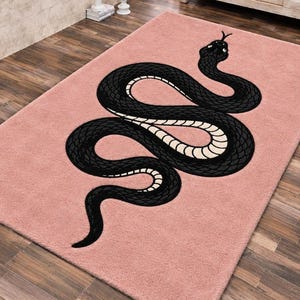 Può includere: Un tappeto rettangolare di colore rosa tenue con una grande grafica centrale raffigurante un serpente nero con un motivo color crema sul ventre. Il serpente è raffigurato in una posizione avvolta a forma di S, che va dall'alto al basso.