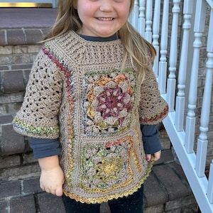 May include: A tan crochet top with colorful floral designs. The top has a square pattern with a round floral design in the center. The sleeves are short and flared. The top is worn over a long-sleeved blue shirt and black pants.
