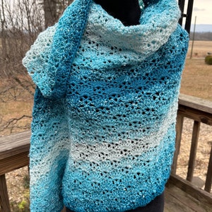 May include: A crocheted shawl in shades of blue and white, with a gradient pattern. The shawl is draped over a black mannequin and is made with a textured yarn.