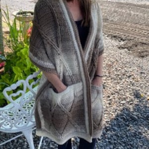 以下が含まれることがあります： 茶色と白の2つのポケットが付いたクロシェのショール。ショールはケーブル編みのパターンとフリンジの縁取りが施されています。