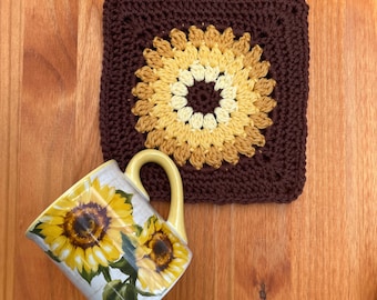 May include: A square crochet coaster featuring a dark brown border and a central sunflower motif in shades of yellow and cream, paired with a white ceramic mug decorated with a yellow sunflower print and a solid yellow handle.