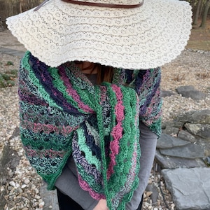 May include: A crocheted shawl with a green, pink, and purple color scheme. The shawl is draped over a person's shoulders and is partially covering a white crochet hat.