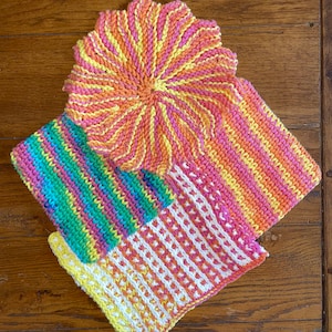 May include: A collection of colorful, handmade knitted washcloths and a circular pot holder. The washcloths feature vibrant stripes and patterns in shades of yellow, pink, orange, and green. The pot holder has a flower-like design with a spiral pattern.