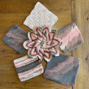 May include: A collection of handmade crochet items, including square and flower-shaped pot holders and washcloths. The color palette features shades of pink, brown, and gray. The items are arranged on a wooden surface, showcasing the texture and craftsmanship.