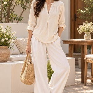 May include: A beige long-sleeved blouse with a split neckline and pleated front detail paired with white wide-leg trousers. The outfit is accessorized with a woven straw tote bag and brown crisscross strap sandals, creating a neutral ensemble.