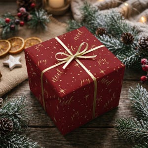 May include: A square gift box wrapped in deep red paper featuring a gold foil pattern of the words joy and noel, interspersed with small stars and dots. The box is tied with a thin, shimmering gold ribbon arranged in a centered bow.
