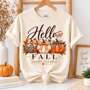 May include: A cream-colored t-shirt featuring the text Hello FALL Sweater Weather in brown script and block letters. The graphic includes three pumpkins decorated with bows, one with a leopard print, one with orange plaid, and one with a solid orange finish.