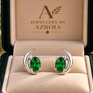 May include: A pair of silver-toned stud earrings featuring oval-cut green gemstones nestled within a crescent moon frame. The earrings are displayed in a dark green velvet jewelry box with the text Jewellery by Azroia printed inside.