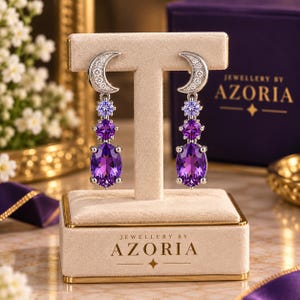May include: A pair of silver-toned drop earrings featuring a crescent moon top encrusted with small clear stones, followed by a light purple round stone, a medium purple round stone, and a large oval deep purple stone at the base.