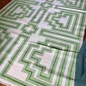 Könnte beinhalten: Ein rechteckiger Teppich mit einem sich wiederholenden geometrischen Muster in leuchtendem Grün und Off-White. Das Design besteht aus ineinandergreifenden, treppenartigen Linien und kreuzförmigen Motiven in einem symmetrischen Raster.