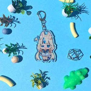 Pode incluir: Um porta-chaves de acrílico transparente com uma personagem de anime de desenho animado com cabelo loiro e um vestido azul e branco. O porta-chaves está preso a um porta-chaves de metal prateado. A imagem é rodeada por pequenas plantas decorativas, um jacaré verde e outros pequenos objetos sobre um fundo azul.