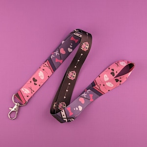 Puede incluir: Un cordón morado y rosa con un patrón repetido de corazones, lazos y nubes. El cordón tiene un clip de metal plateado.