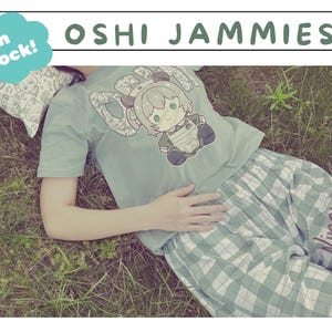 May include: A person wearing a light green t-shirt and plaid shorts, lying on grass. The t-shirt features a cartoon character design and the text "OSHI JAMMIES". The shorts have the text "Nimi Nightmare". A pillow is visible.