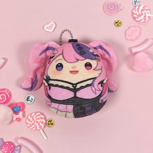 Puede incluir: Un llavero de peluche rosa y morado con un personaje de dibujos animados con cabello rosa y un atuendo blanco y negro. El personaje tiene una gran sonrisa y lleva un collar negro con un colgante de corazón. El llavero está rodeado de caramelos rosas y blancos.