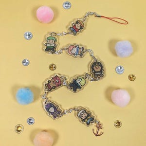May include: A clear keychain with a silver chain and charms featuring cartoon characters from the anime One Piece. The charms are round and feature different characters from the show, including Luffy, Zoro, and Sanji. The keychain also has a small anchor charm at the end.