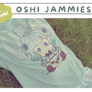Può includere: T-shirt azzurra con un personaggio anime dei cartoni animati. La maglietta presenta la scritta "OSHI JAMMIES" e "In Stock!" in una nuvola verde. L'indumento è indossato su una superficie erbosa.