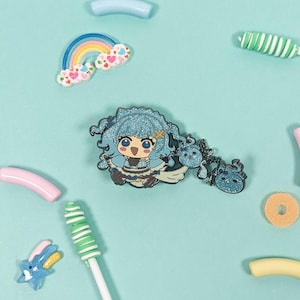 May include: An assortment of colorful, whimsical accessories on a light blue background. The image features a glittery anime-style character pin, a rainbow, lollipops, and various candy-shaped charms. The items are arranged in a playful, decorative manner.