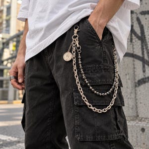 May include: A silver-toned multi-strand wallet chain featuring a thick curb chain, a secondary strand of faceted dark beads, and a circular coin pendant with a profile portrait. The chain attaches to the belt loop of black cargo pants.