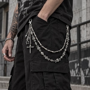 May include: A silver-toned metal wallet chain featuring a double-layered design with skull and bone-shaped links. The chain includes a dangling charm cluster with a skeleton figure, small skulls, and a cross-shaped pendant attached by a lobster clasp.