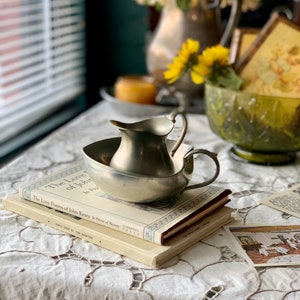 Op de afbeelding: Een zilveren roomkan en suikerpot set op een stapel boeken met een wit kant tafelkleed. De boeken zijn getiteld "The Love Poems of John Keats is Praise of Beauty".