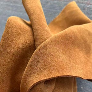 May include: A piece of tan suede leather with a soft, matte, and slightly fuzzy texture. The material is folded, creating deep curves and shadows that highlight the consistent light brown color and the natural, fibrous surface of the hide.