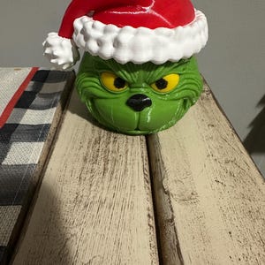 Puede incluir: Una cabeza de personaje esculpida, redonda y verde, con el ceño fruncido, ojos amarillos y nariz negra, que lleva un gorro de Papá Noel rojo brillante con un borde blanco texturizado y un pompón blanco, con acabado de impresión 3D visible.