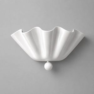 Może przedstawiać: Biały kinkiet ceramiczny z szerokim, półkolistym kloszem o głębokich, pionowych plisach tworzących falisty brzeg. Pojedyncza, gładka biała kula jest przymocowana do środkowej dolnej części oprawy, stanowiąc centralny punkt dekoracyjny.