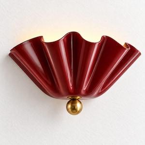 Può includere: Una lampada da parete con un paralume a forma di ventaglio di colore rosso intenso, lucido, con un bordo plissettato e ondulato. L'apparecchio è fissato alla base da un elemento decorativo sferico in metallo color ottone.
