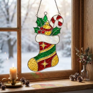 Peut inclure: Un attrape-soleil en vitrail en forme de chaussette de Noël avec un revers blanc, des rayures diagonales rouges et vertes et des accents jaunes. Il est orné de feuilles de houx vertes, de baies rouges, d'une boule jaune et d'un sucre d'orge.