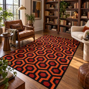 May include: A rectangular area rug featuring a repeating geometric pattern of interlocking orange and black hexagons. The rug is positioned on a wooden floor in a room with a leather armchair, a bookshelf, and a cream-coloured accent chair.
