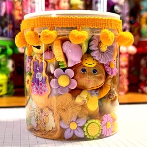 May include: A clear plastic cylindrical container filled with assorted colorful plastic charms, including a yellow doll figure, purple and green flowers, and a purple bear on a cupcake. The lid is decorated with a bright yellow pom-pom trim.