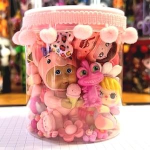 Puede incluir: Un recipiente cilíndrico de plástico transparente lleno de varias figuras pequeñas de plástico de colores, incluyendo un personaje con disfraz de cerdo sosteniendo un helado, una criatura rosa tipo lagarto y una muñeca rubia, con un borde de pompones rosas.