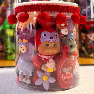 May include: A clear plastic cylindrical container filled with various miniature toys, including a doll in a red monkey costume holding a cupcake, a miniature Hershey's syrup bottle, and small animal figures. The lid is trimmed with red pom-pom fringe.
