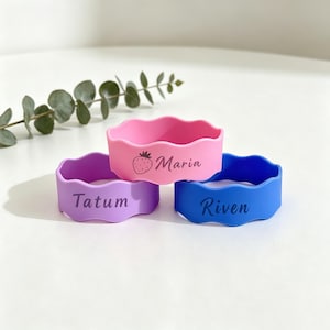 May include: Three wavy silicone bracelets in pink, purple, and blue. The pink bracelet features a strawberry icon and the name Maria. The purple bracelet is printed with the name Tatum, and the blue bracelet is printed with the name Riven.