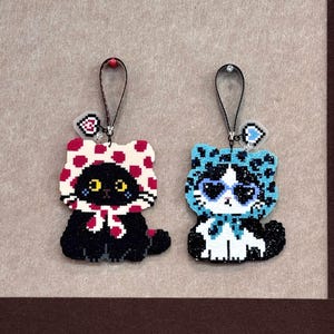 May include: Two pixel art cat ornaments with black hanging loops. The left cat is black with a red-spotted white hood and a small heart charm. The right cat is black and white with a blue leopard-print hood, heart-shaped sunglasses, and a heart charm.