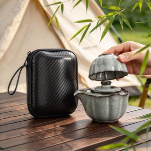 Puede incluir: Un juego de té portátil que incluye una tetera de cerámica gris claro con acabado de esmalte craquelado y tapa a juego, acompañado de una pequeña taza de cerámica y un estuche de transporte negro con textura de fibra de carbono.
