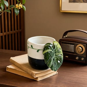 May include: A ceramic mug featuring a two-tone design with a white upper section and a black lower section. A three-dimensional, glossy green monstera leaf is attached to the side of the mug, and a thin green vine pattern encircles the white rim.