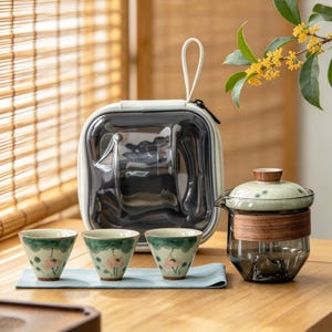May include: A tea set consisting of three small ceramic cups with hand-painted pink lotus flowers and green leaves, alongside a glass teapot with a wooden band, a light green lid, and a matching white carrying case with a clear front panel.