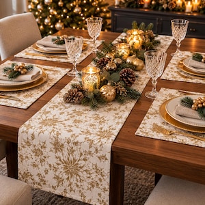 May include: A dining table set with a white and gold snowflake patterned runner and matching placemats. Each place setting features a white plate, a gold charger, a beige napkin topped with a gold berry and pine sprig, and a crystal glass.