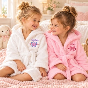 May include: Two plush hooded robes, one white and one pink, featuring long sleeves and a tie waist. The white robe is embroidered with the name Sophia and a blue butterfly. The pink robe is embroidered with the name Mila and a pink bow.