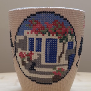 Pode incluir: Um copo cilíndrico com um design de pintura de diamante no exterior. A arte retrata um edifício branco com portas e janelas azuis, acentuado por flores vermelhas contra um céu azul, tudo composto por pequenas contas de plástico coloridas.