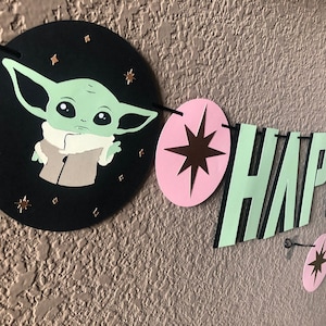 Pode incluir: Uma faixa de papel verde e preta com as palavras "HAPPY" em letras verdes com contornos pretos. A faixa tamb&eacute;m apresenta um c&iacute;rculo rosa com uma estrela marrom e um c&iacute;rculo preto com um personagem verde da franquia Star Wars.