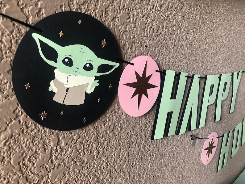 Pode incluir: Uma faixa de papel verde e preta com as palavras "HAPPY" em letras verdes com contornos pretos. A faixa tamb&eacute;m apresenta um c&iacute;rculo rosa com uma estrela marrom e um c&iacute;rculo preto com um personagem verde da franquia Star Wars.