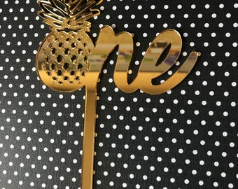 May include: Gold mirror acrylic cake topper with the word "one" and a pineapple design.