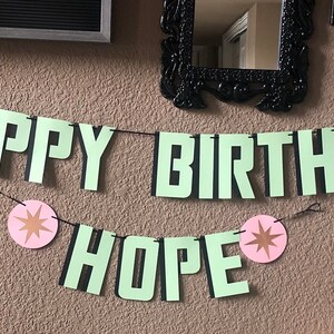 Pode incluir: Uma faixa verde e preta com as palavras "Happy Birthday Hope" e decora&ccedil;&otilde;es de estrelas rosa e douradas. A faixa apresenta um personagem de desenho animado da franquia Star Wars.
