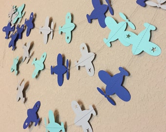 May include: A paper airplane garland with blue, gray, and light blue airplanes. The airplanes have a star cut out of the center. The garland is strung together with clear thread.